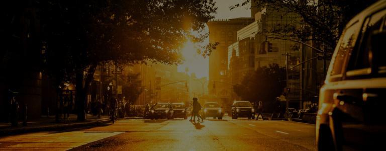 Menschen, die bei Sonnenuntergang eine verkehrsreiche Straße überqueren. Das Bild enthält viel Orange und Gelb – Aufnahme von der Mitte der Straße.